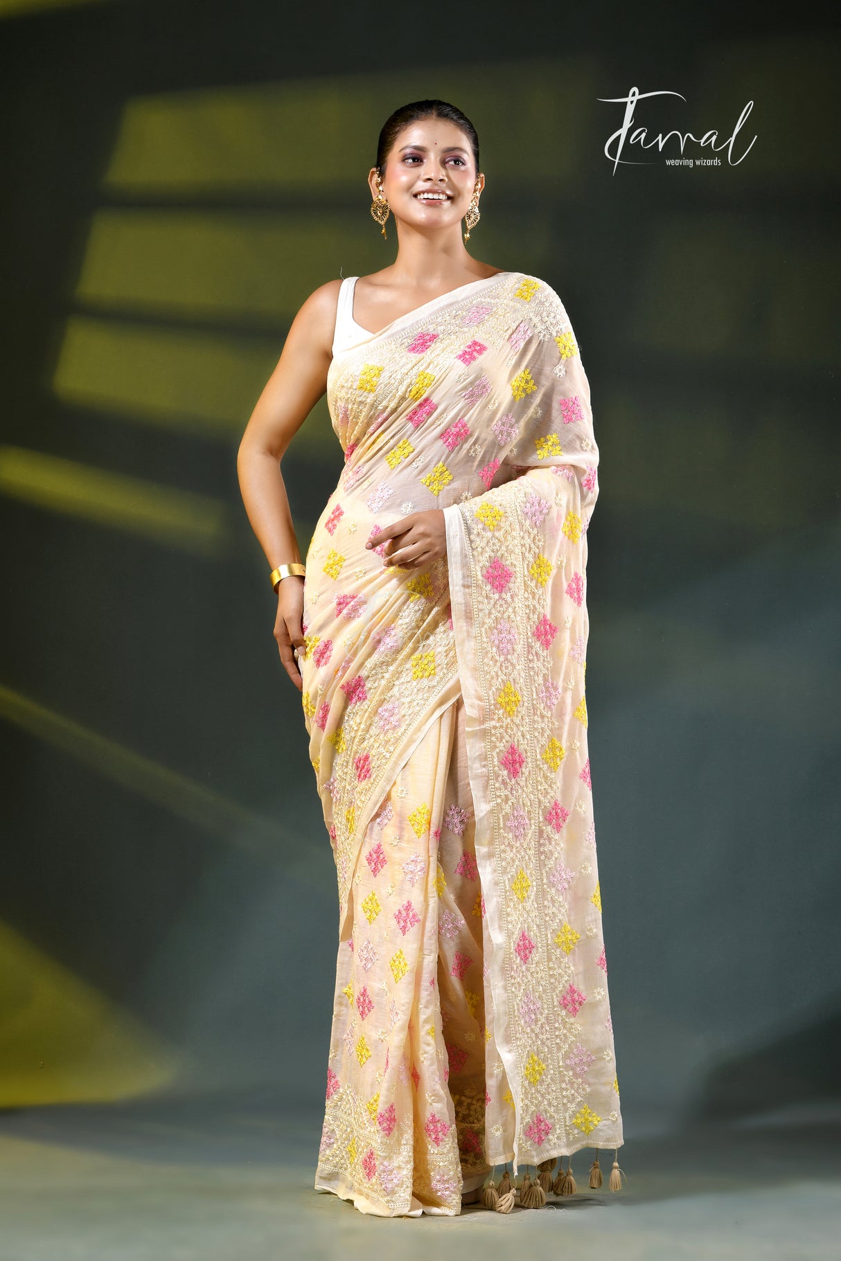 A woman modeling a light beige saree with pink and yellow floral patterns, standing on a dark stage with a 'Farral' watermark in the upper right.