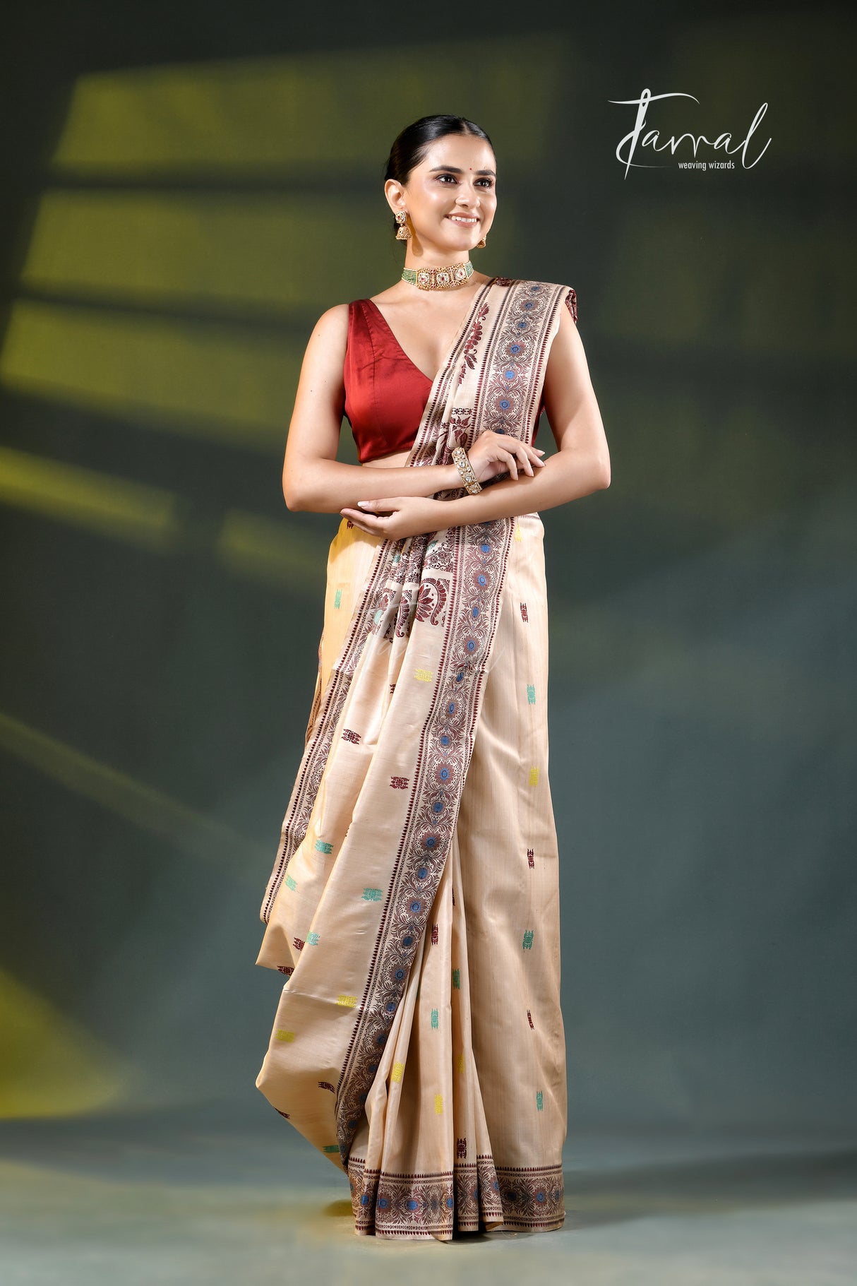 A woman modeling a beige saree with a red sleeveless blouse, standing against a dark stage-like background with yellow lighting. The saree has a patterned border in blue, brown, and gold, and the image features a 'Farral' brand watermark in the upper right.