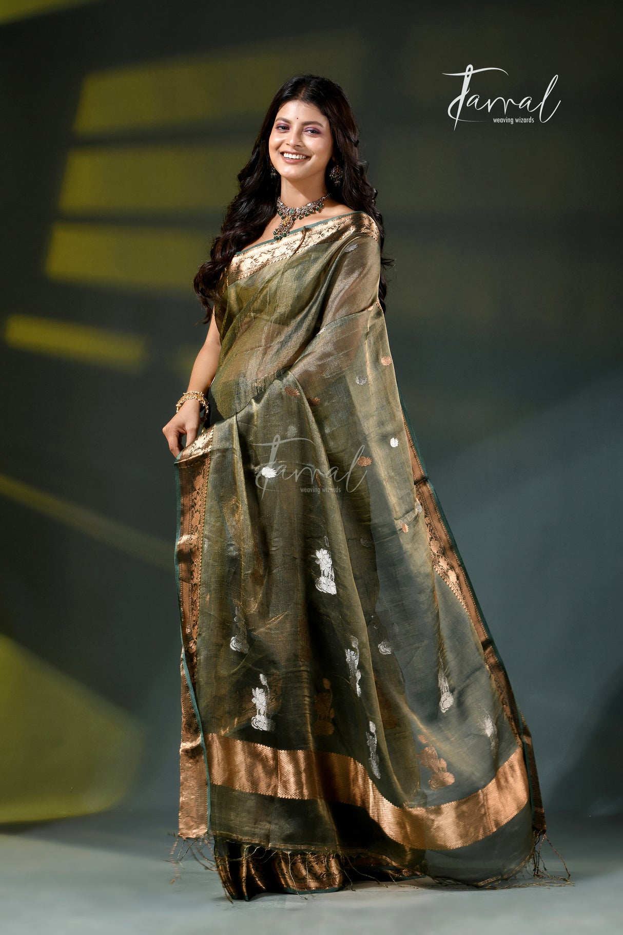 A woman modeling a dark olive green saree with metallic gold borders and silver motifs, standing on a runway under a dark stage backdrop with yellow lighting accents. The brand name 'Jarral' is visible as a watermark and logo.