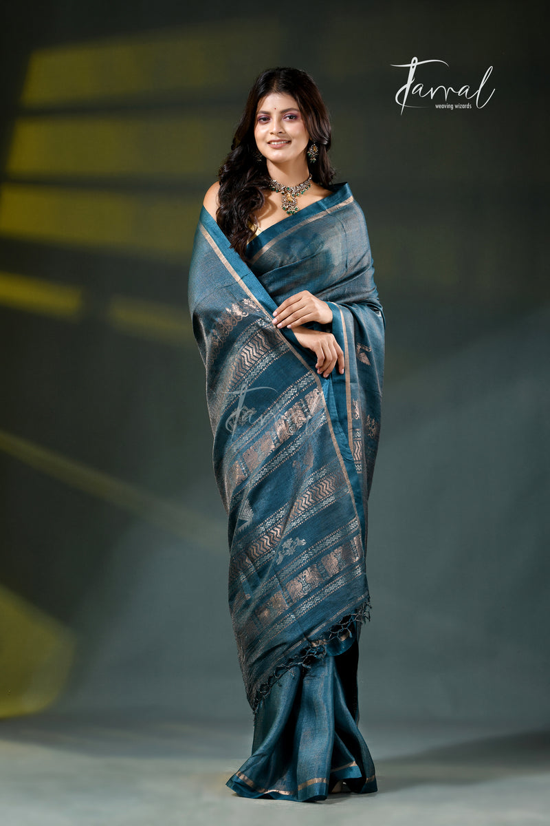 A woman modeling a dark teal blue saree with metallic gold and silver geometric patterns, standing against a dark gray studio backdrop with yellow light beams. The brand name 'Jarral' is visible in the top right corner.