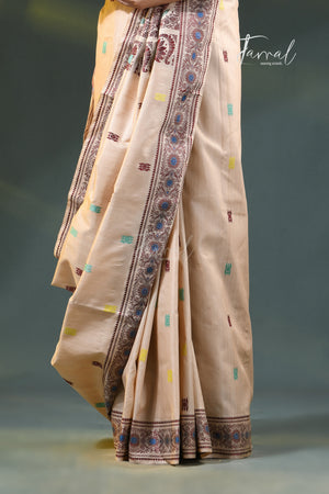 A draped beige saree with a wide brown border featuring intricate geometric and floral patterns, accented with small multicolored motifs, shown against a dark gray studio background.