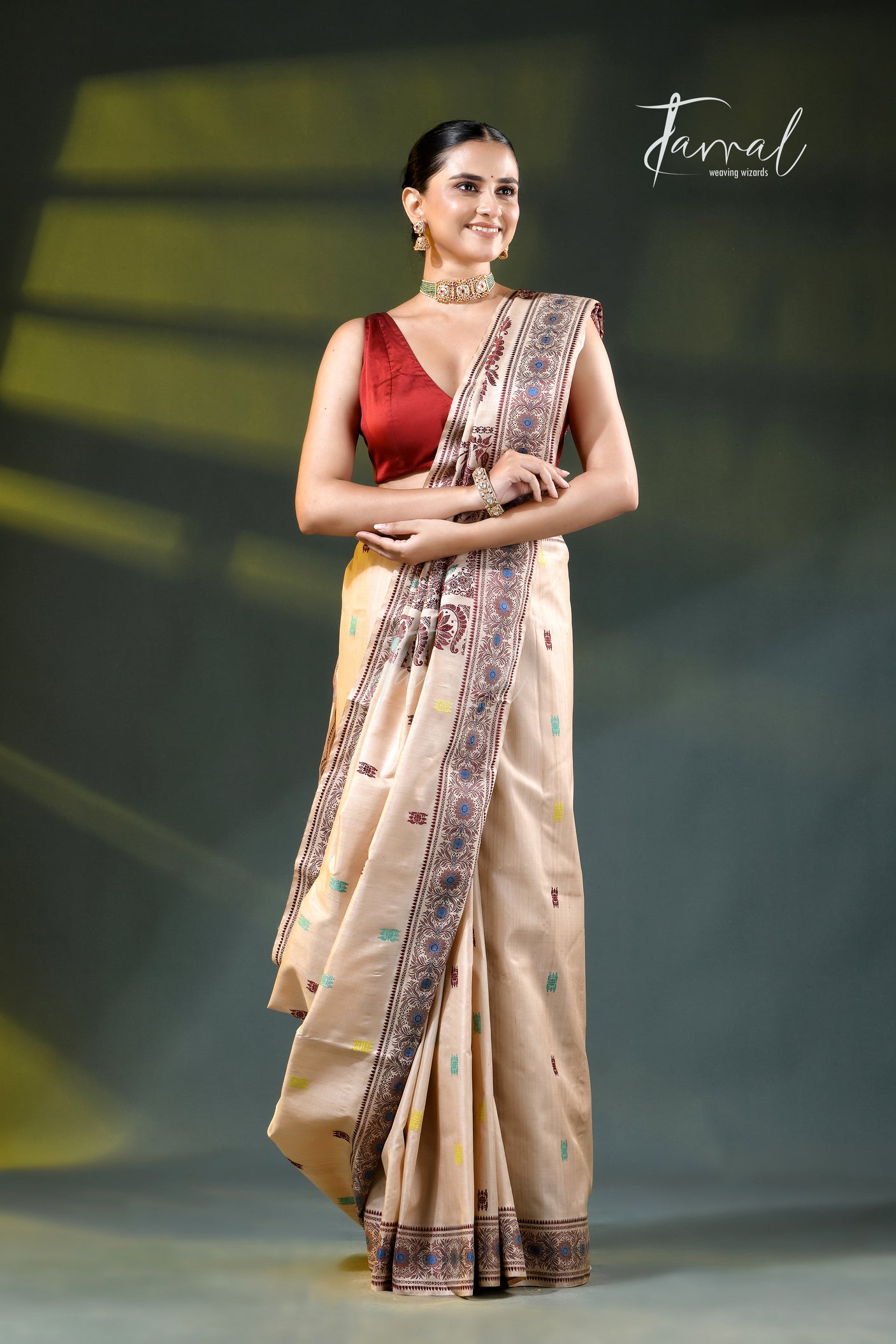 A woman modeling a beige saree with a red sleeveless blouse, standing against a dark stage-like background with yellow lighting. The saree has a patterned border in blue, brown, and gold, and the image features a 'Farral' brand watermark in the upper right.