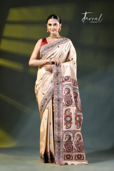 A woman modeling a beige silk saree with intricate multicolor paisley and floral motifs on the border, paired with a red sleeveless blouse. The image is branded with 'Farral' in the top right corner against a dark stage-like background with soft yellow spotlights.