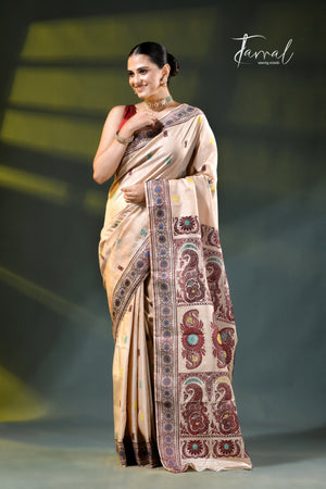 A woman models a beige silk saree with intricate brown, red, and blue paisley and floral motifs, standing against a dark gray stage background with greenish lighting. The brand name 'Jarral' is visible in the top right corner.
