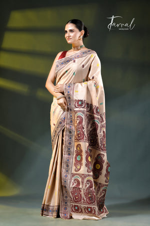 A woman models a beige saree with intricate multicolor paisley and floral patterns, featuring a red blouse underneath. The brand name 'Jarral' is visible in the top right corner against a dark, softly lit background.