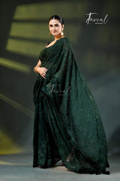 A woman modeling a dark forest green embroidered saree with a matching sleeveless blouse, standing on a dimly lit stage with spotlighting. The brand name 'Farral' is visible as a watermark and logo.