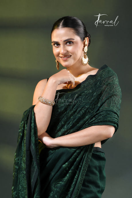 A woman modeling a dark forest green saree with a subtle jacquard pattern, styled with gold jewelry against a dark green studio background. The brand name 'Jarral' is visible as a watermark and logo.