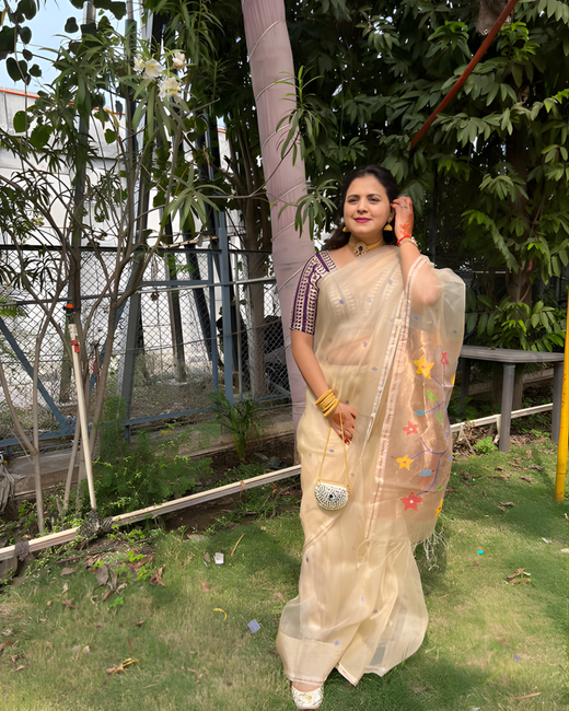 Woman in a light-colored saree standing outdoors with greenery in the background