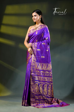 A woman modeling a purple silk saree with intricate gold zari motifs and borders, standing on a stage with a dark background and spotlighting. The brand name 'Jarral' is visible as a watermark and logo.